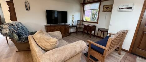 Ein Wohnzimmer mit einem beigen Sofa, einem Holzstuhl mit blauer Kissenauflage, einem Fernseher, einem Holzschrank und einem Fenster mit Jalousien.
