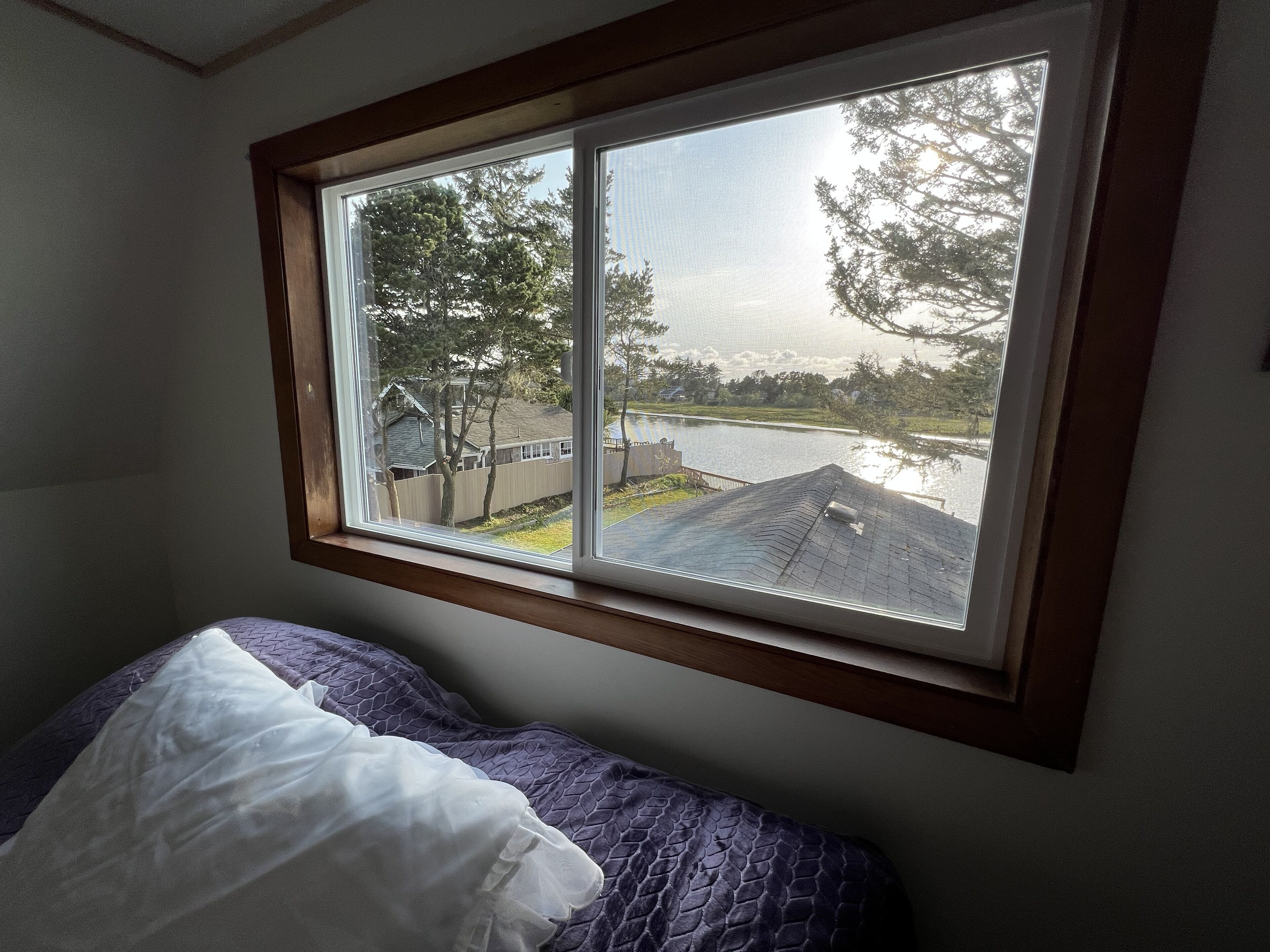 A bedroom with a window overlooking a lake and a house.