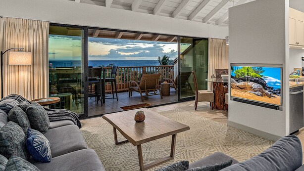 A modern living room with a large sofa, a coffee table, and a television. There is a sliding glass door leading to a balcony with outdoor furniture.