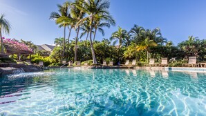 A clear pool with lounge chairs and palm trees, surrounded by lush greenery.