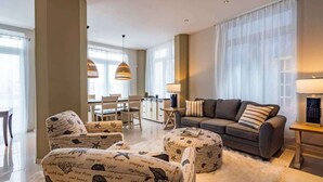 Una sala moderna con un sofá, un reposapiés y sillas estampadas, un comedor con mesa y sillas, y ventanas amplias con cortinas translúcidas.