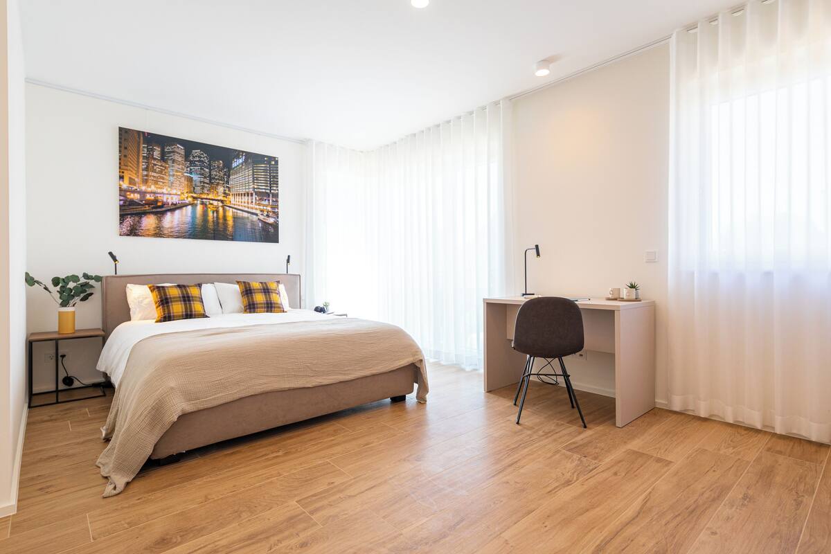 Quarto moderno com uma cama grande, uma escrivaninha e uma pintura da paisagem urbana na parede.