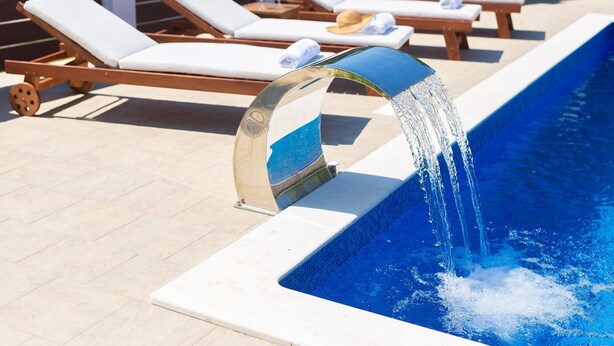 Una piscina con una fuente moderna, rodeada de sillas de descanso y una tumbona.