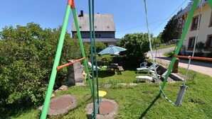 Ein Spielplatz mit Schaukeln und Rutsche, umgeben von Gras und einem Haus im Hintergrund.