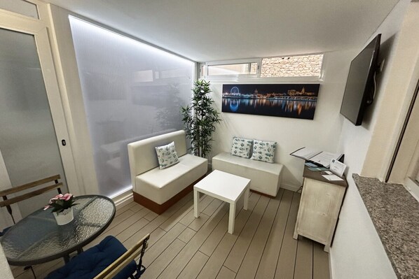 A compact living space with a sofa, a small table, a TV, and a window with a cityscape view. - Interior Studio Lirio - Historical Center (Málaga)