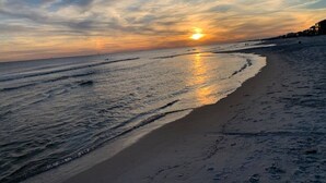 A beach with a sunset over the ocean, sandy shore, and a few buildings in the background. - Ocean+Lake Views~HEATED Pool~Pickleball~Golf Cart (Gulf Shores)
