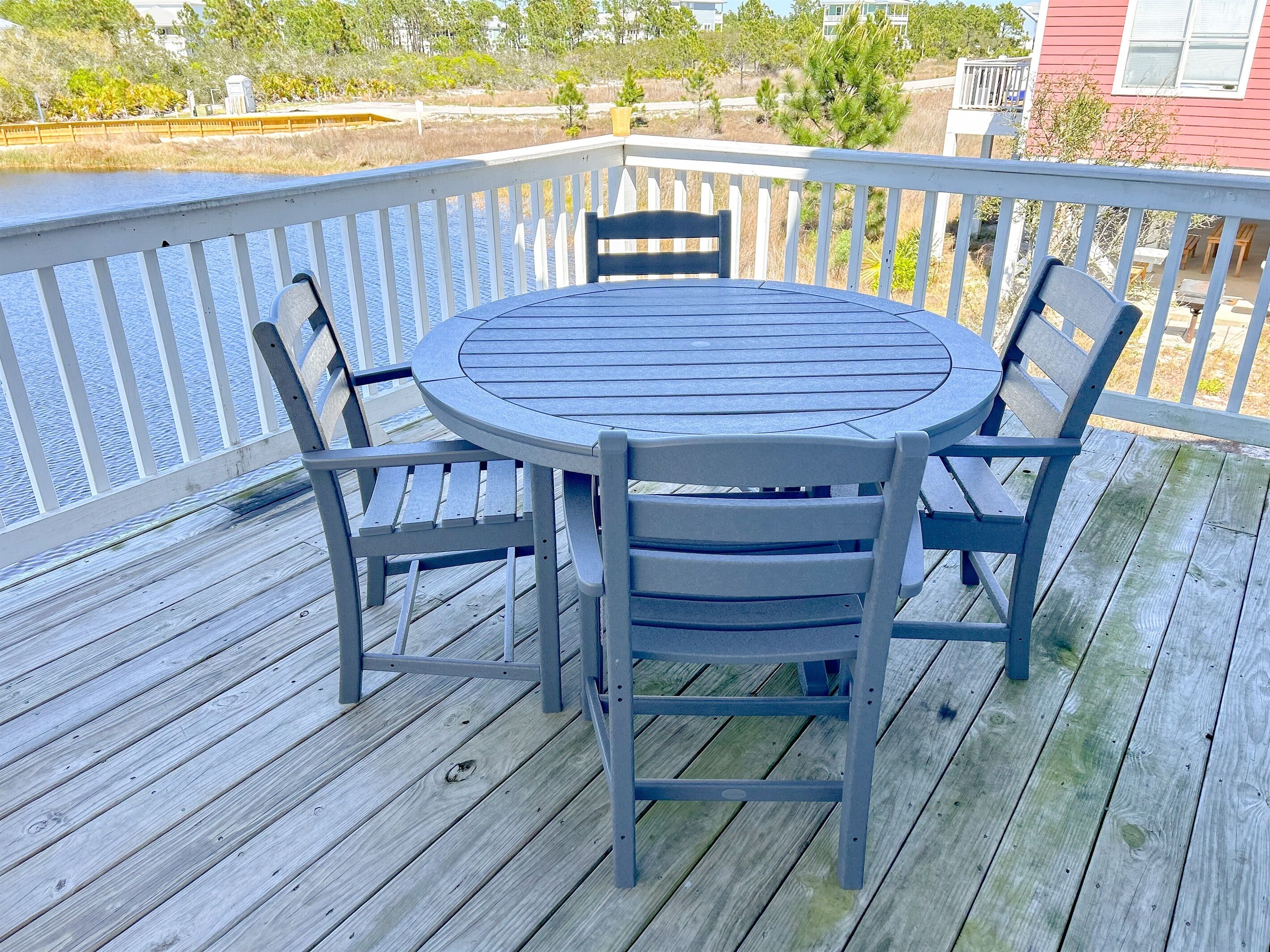 A round table with four chairs on a wooden deck with a railing, overlooking a body of water and greenery.