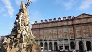 A classical fountain with a statue of a figure ascending a rocky structure, set against a backdrop of a large, ornate building with multiple arches and a courtyard.