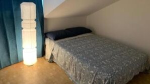 A small, dimly lit room with a single bed, a nightstand, and a tall, white lamp.