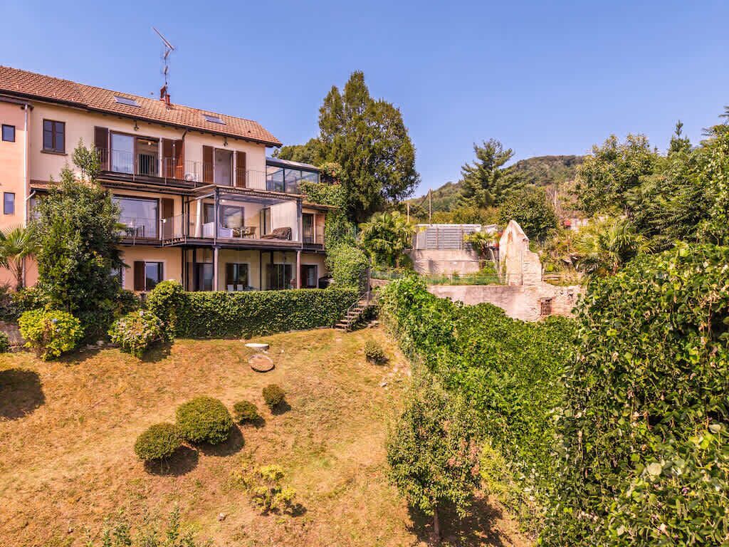 Ein zweistöckiges Haus mit Balkon, ein Garten mit Büschen und einer kleinen Rasenfläche, sowie ein Steinbau im Hintergrund.