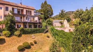 Ein zweistöckiges Haus mit Balkon, ein Garten mit Büschen und einer kleinen Rasenfläche, sowie ein Steinbau im Hintergrund.