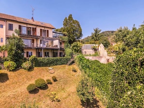 A two-story house with a balcony, a garden with bushes and a small lawn, and a stone structure in the background.