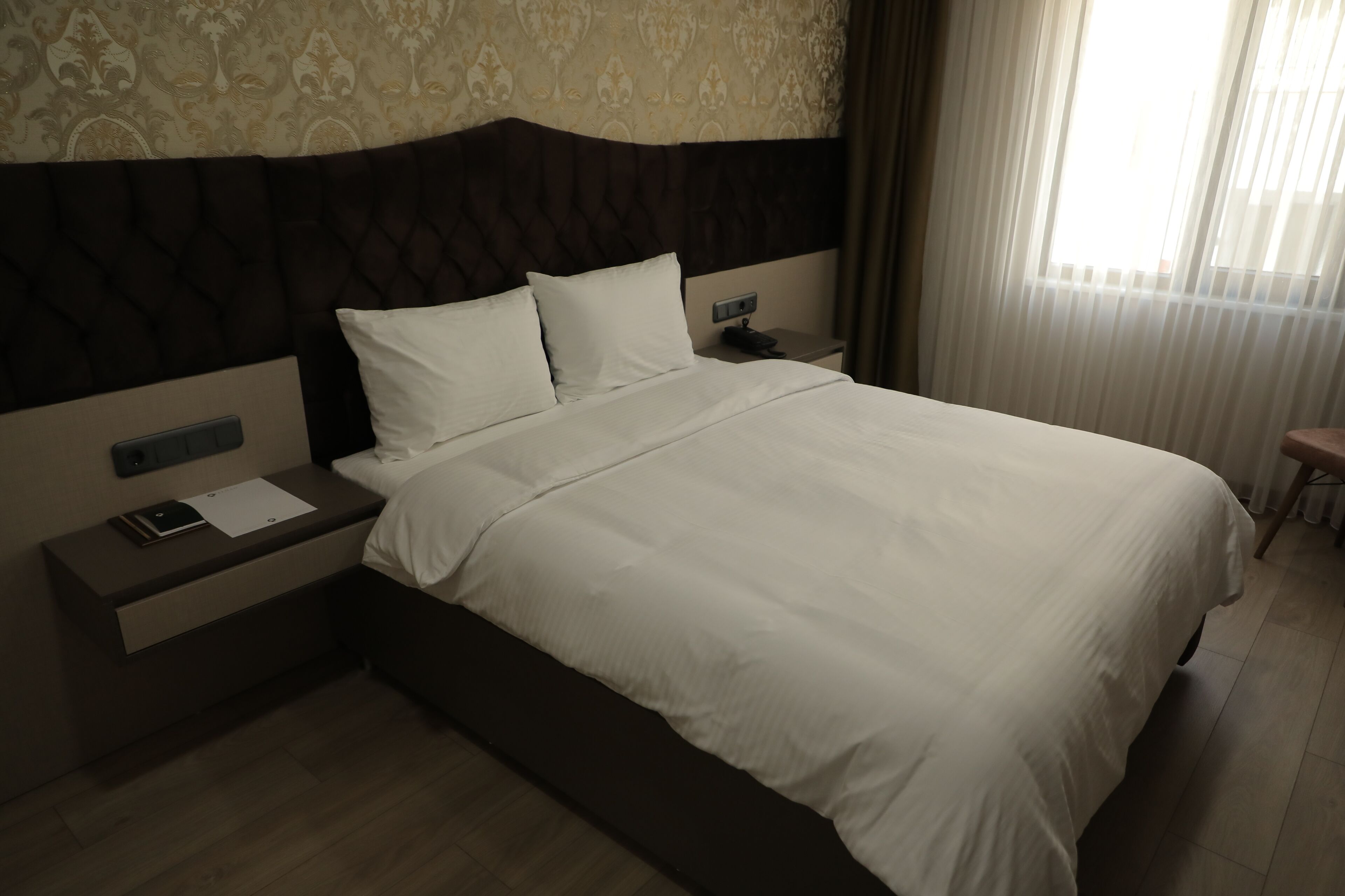 A hotel room with a large bed, a nightstand with a phone and papers, and a window with curtains.