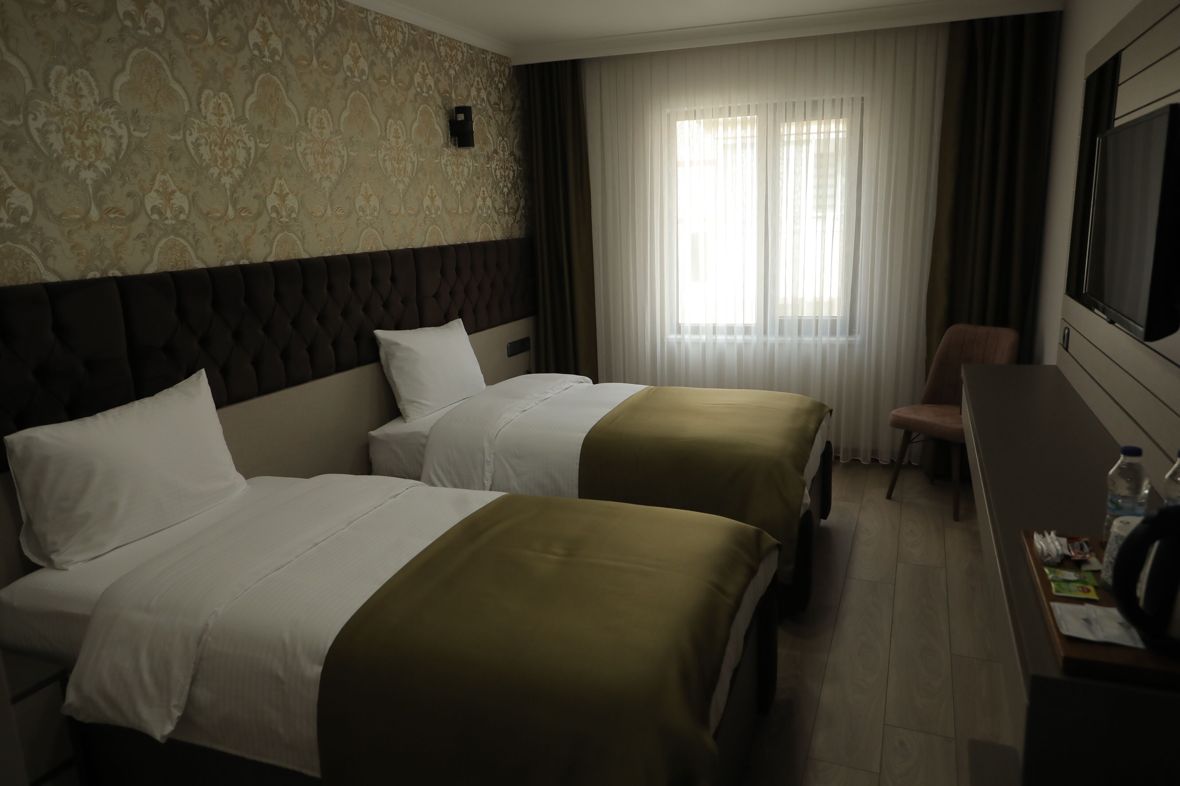 A hotel room with two beds, a desk, a chair, and a large window with curtains.