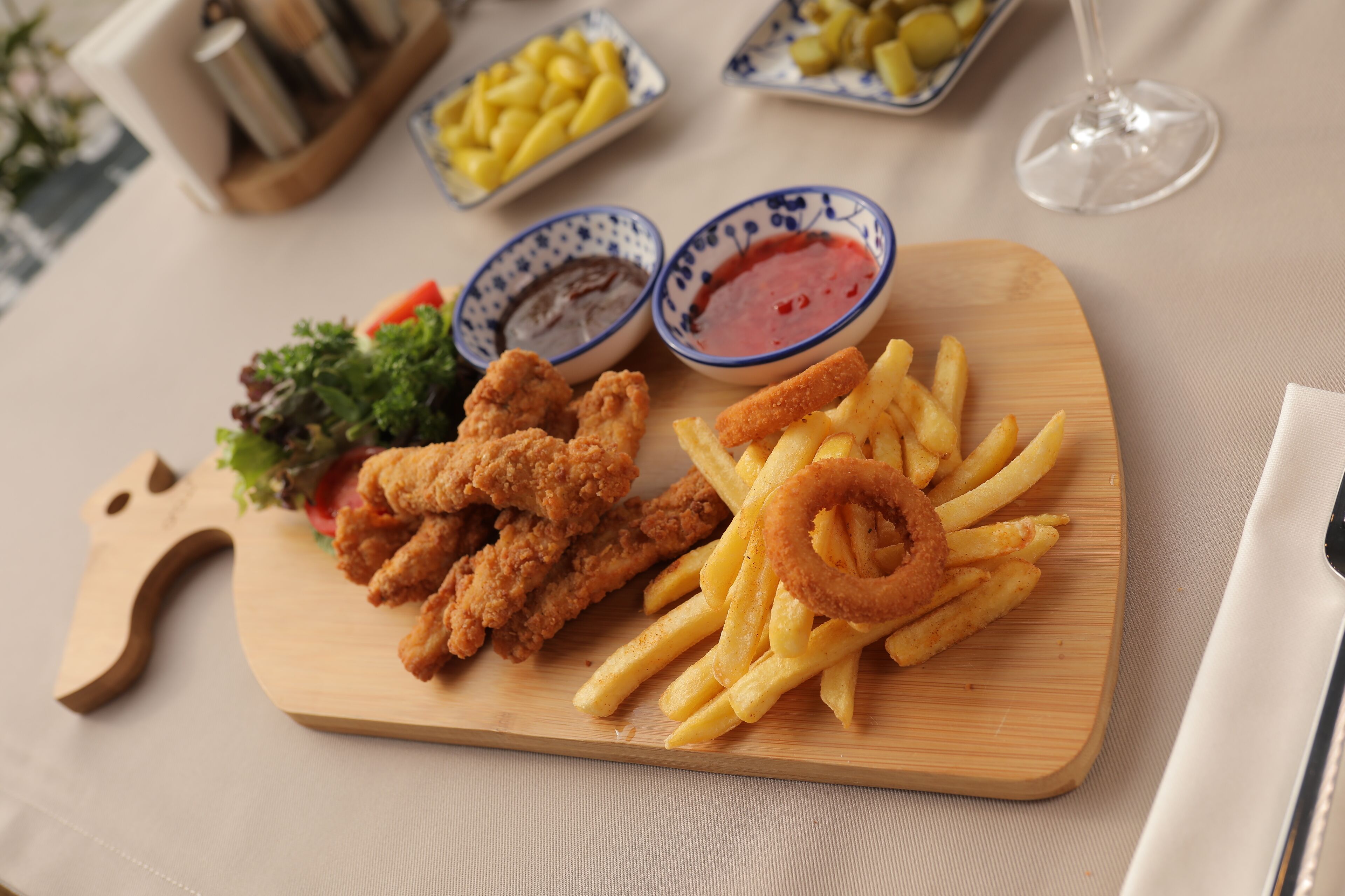 A wooden board with fried chicken, onion rings, and French fries, accompanied by two small bowls of dipping sauces.