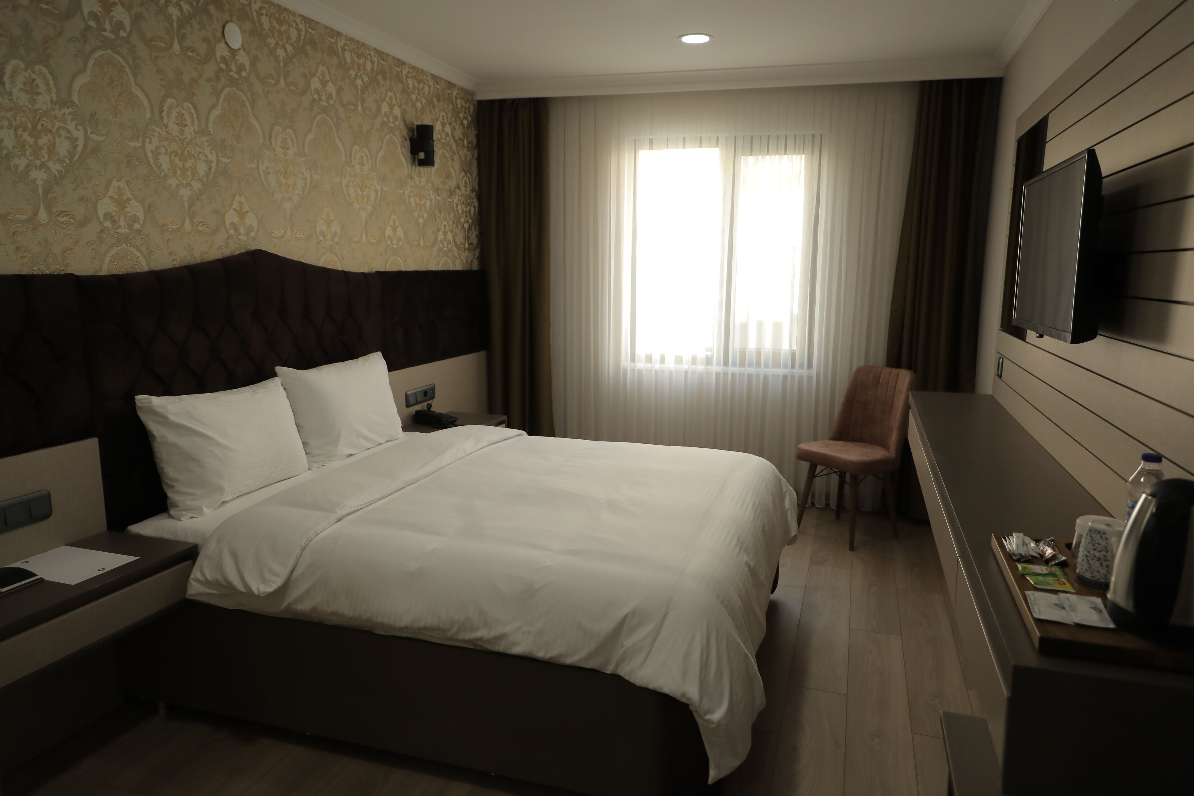 A hotel room with a large bed, a flat-screen TV mounted on the wall, a chair, and a desk with a kettle and a bottle.