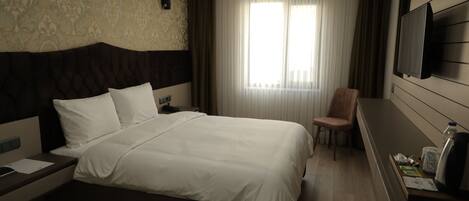 A hotel room with a large bed, a flat-screen TV mounted on the wall, a chair, and a desk with a kettle and a bottle.