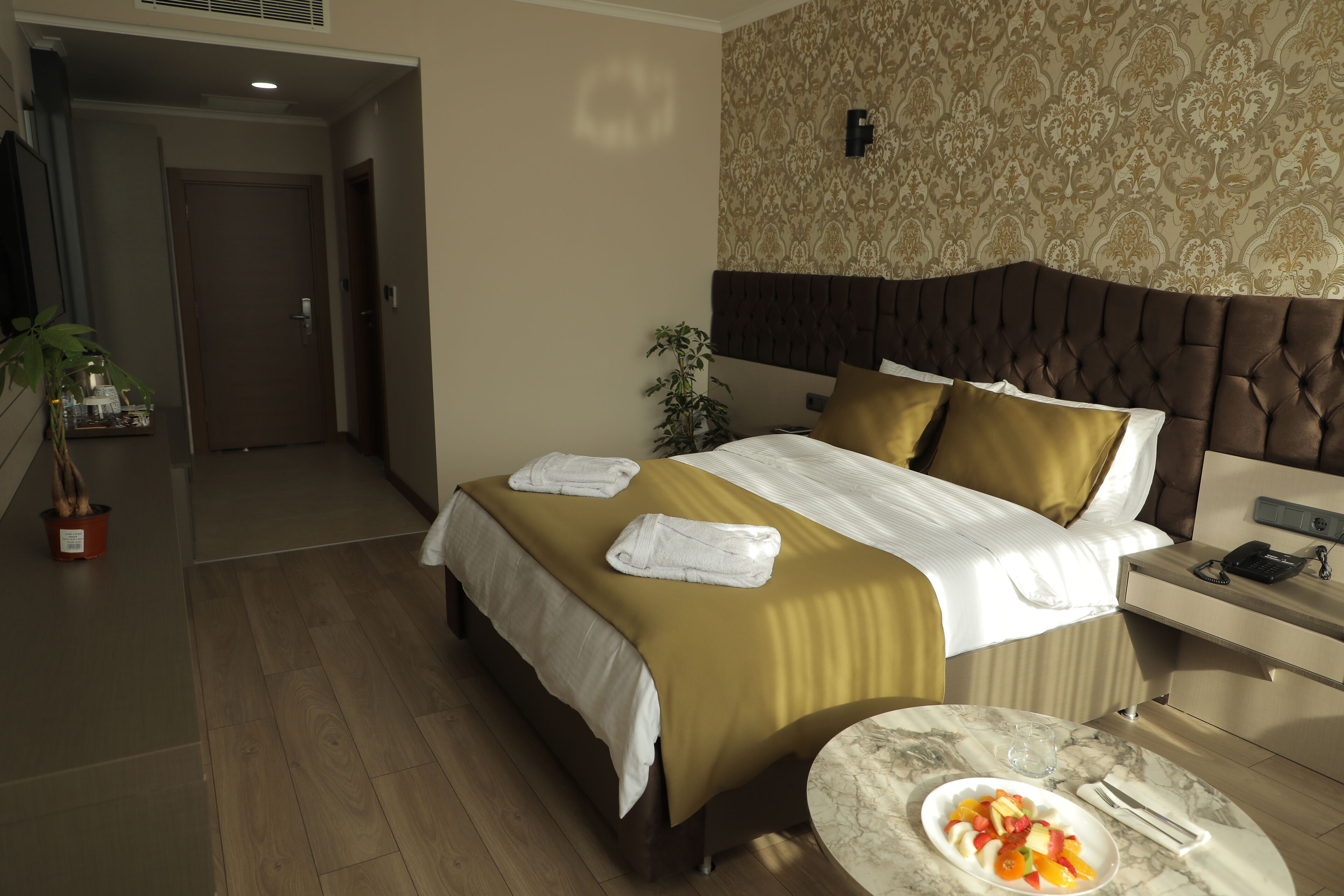 Une chambre d’hôtel avec un lit, une table de chevet sur laquelle se trouve un panier de fruits, une plante en pot et un mur recouvert d’un papier peint à motifs.