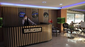 A reception desk with a 'RECEPTION' sign, two clocks, and a potted plant. - ELİT OTELHAN (Amasya)