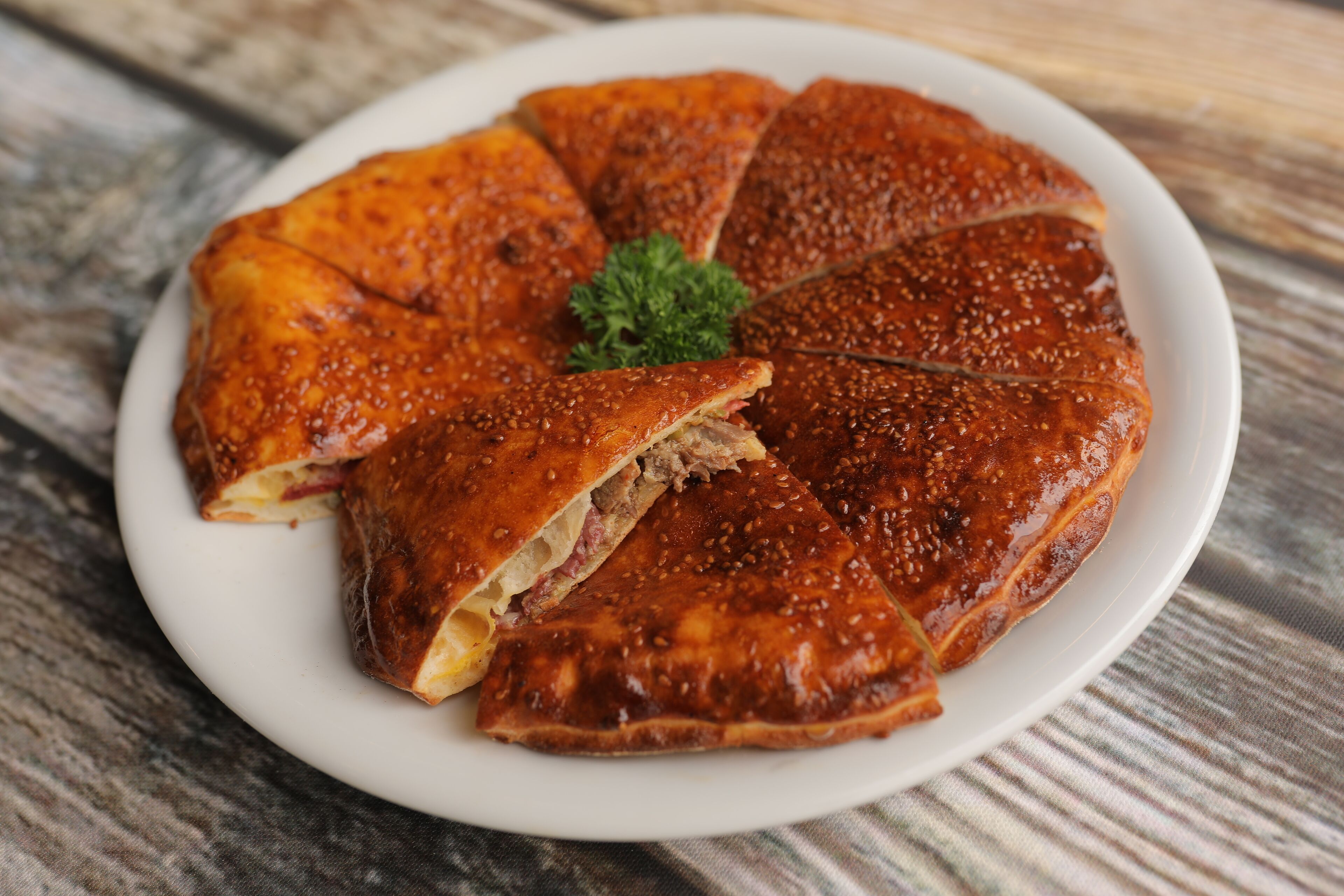 Um pastel redondo e dourado, coberto com sementes de sésamo, sobre um prato branco, decorado com salsa.
