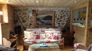 A rustic room with a stone wall, wooden furniture, a sofa with patterned cushions, and a painting of a mountain scene.