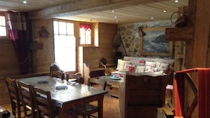 A rustic dining area with a wooden table and chairs, a stone wall with a painting, and a cozy seating area with cushions and a red towel.