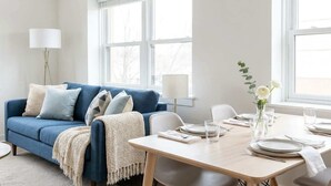 A modern living room with a blue sofa, white and blue cushions, a white table set for dining, and a vase with white flowers. - Central Dudley 1BR Flat w/ Fast Wi-Fi (Dudley)