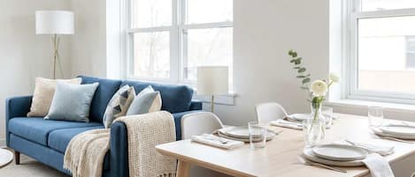 A modern living room with a blue sofa, white and blue cushions, a white table set for dining, and a vase with white flowers.