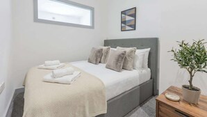 A bedroom with a bed, a window, a wall-mounted picture, a bedside table with a plant, and a potted plant on the floor. - Central Dudley 1BR Flat w/ Fast Wi-Fi (Dudley)