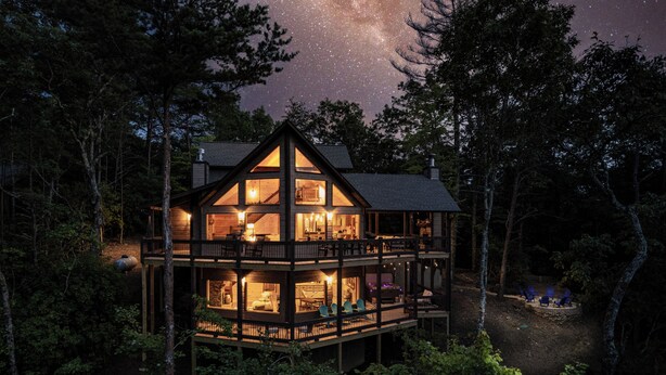 A multi-story wooden cabin with a balcony, surrounded by trees, and a starry night sky above.