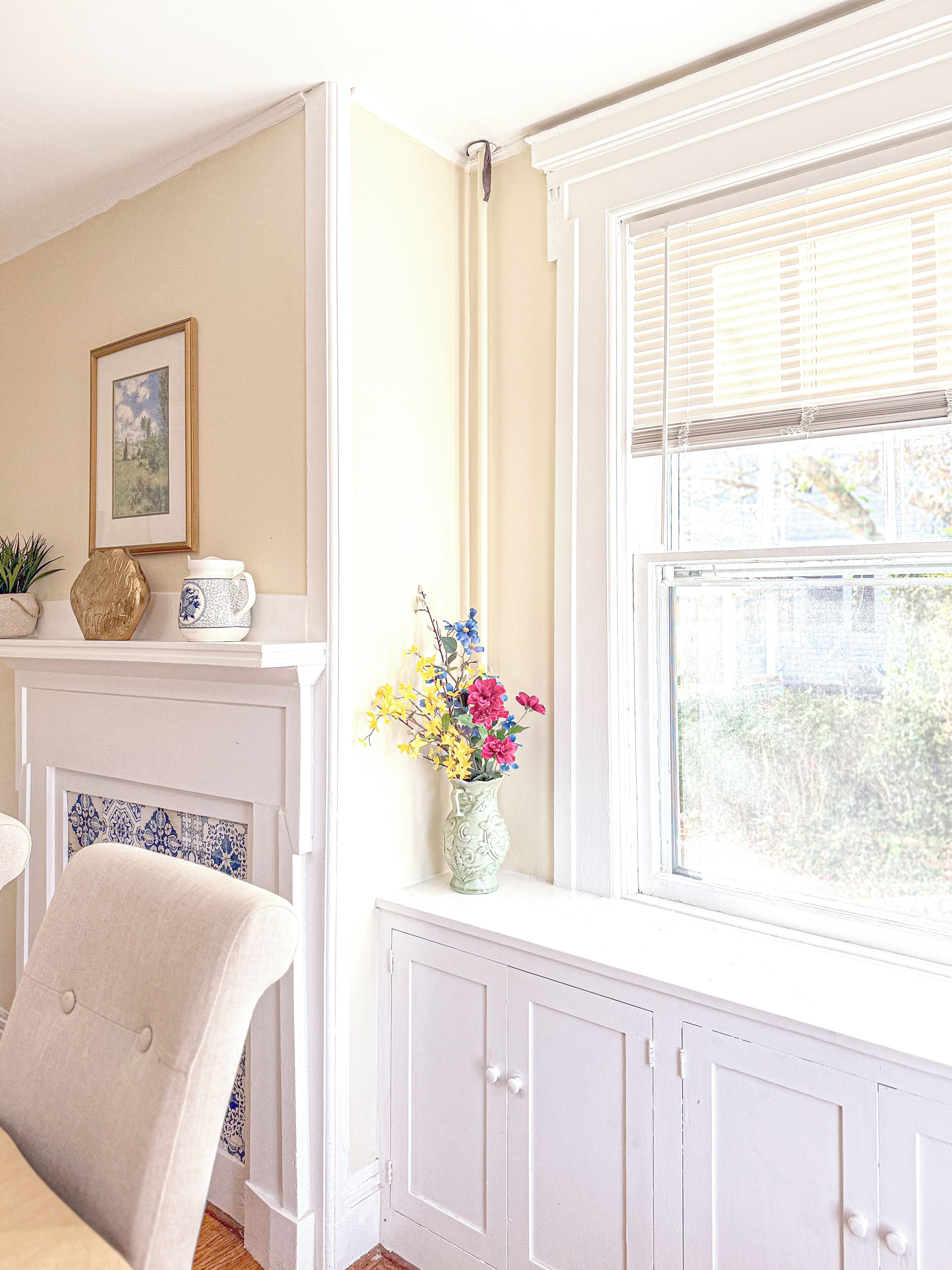 A room with a fireplace, a window with blinds, a vase with flowers, and a framed picture on the wall.