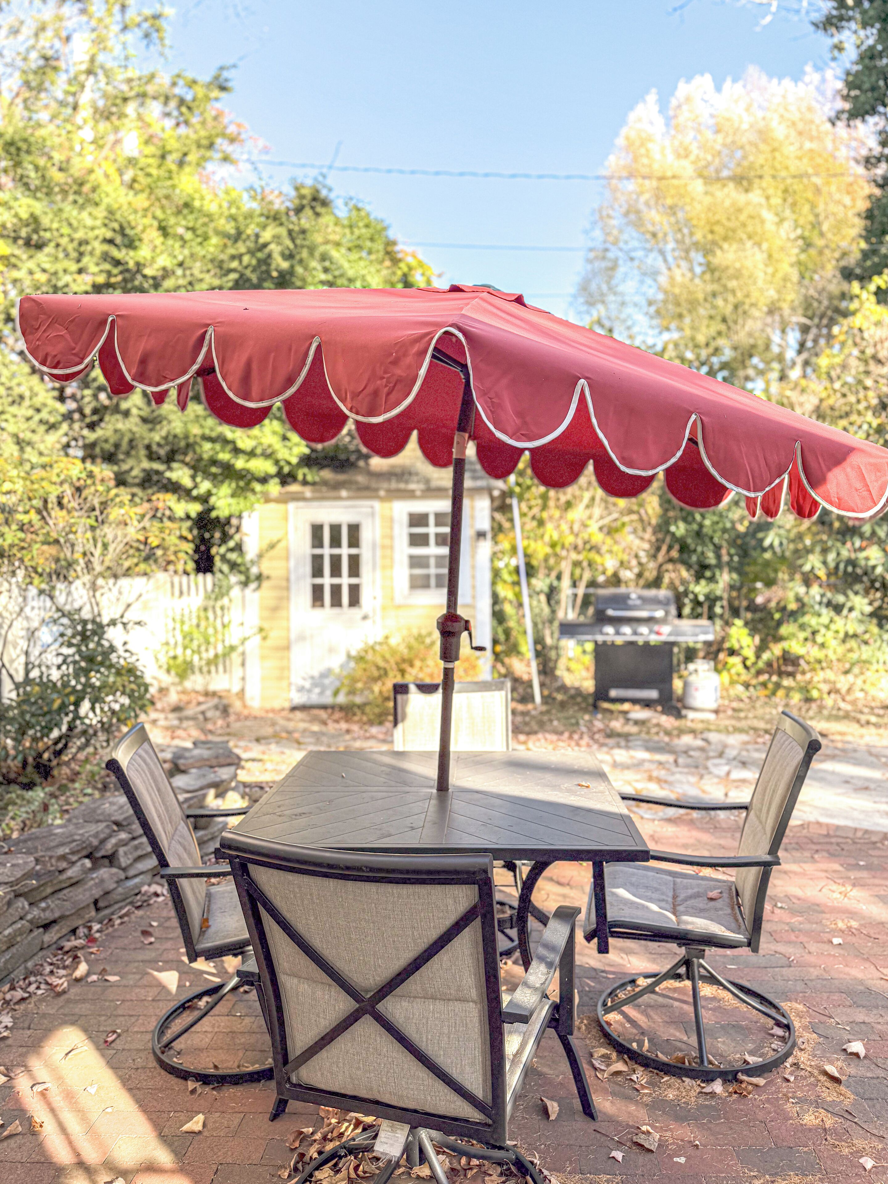 Une terrasse avec une table et des chaises sous un parasol rouge, entourée d’arbres et d’une maison.