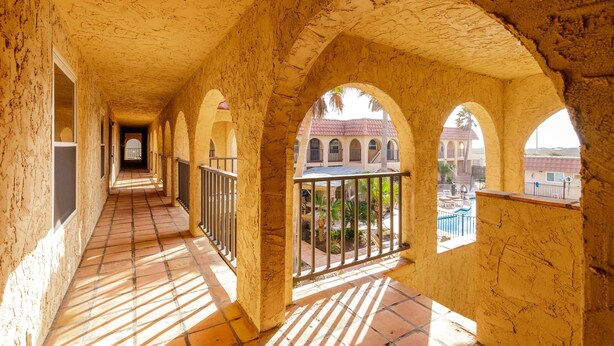 A corridor with arches and a tiled floor, leading to a balcony with a view of a pool and buildings. - Beachside Cozy Condo | Tropical Pool & Coastal Comfort (Corpus Christi)
