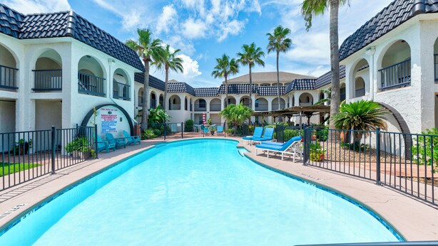 A swimming pool surrounded by lounge chairs and palm trees, with a building featuring arched windows and a tiled roof in the background. - Beachside Cozy Condo | Tropical Pool & Coastal Comfort (Corpus Christi)