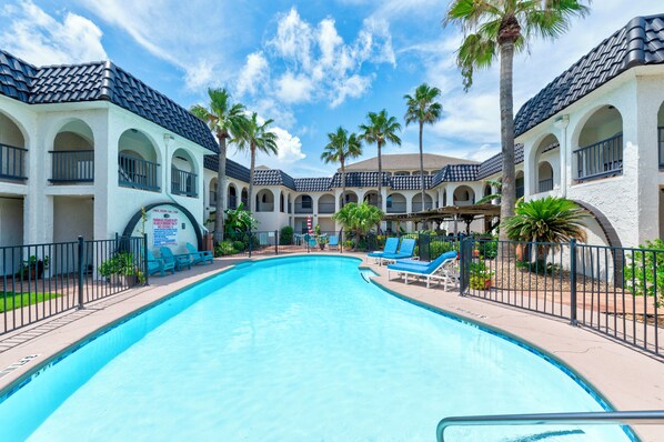 A swimming pool surrounded by lounge chairs and palm trees, with a building featuring arched windows and a tiled roof in the background. - Beachside Cozy Condo | Tropical Pool & Coastal Comfort (Corpus Christi)
