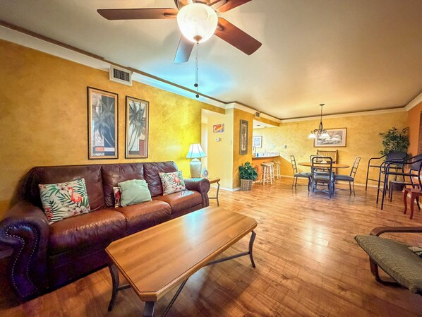 A living room with a brown leather sofa, a wooden coffee table, and a ceiling fan. - Beachside Cozy Condo | Tropical Pool & Coastal Comfort (Corpus Christi)