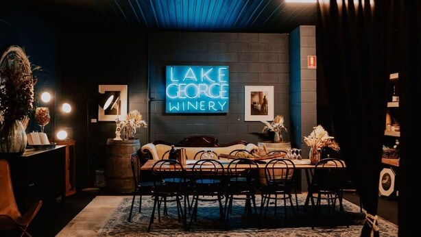 A dimly lit room with a neon sign reading 'LAKE GEORGE WINERY,' a wooden table with chairs, and a sofa. - Baxter at Lake George Winery by Tiny Away (Lake George)