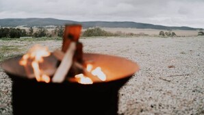 A campfire with flames, set on a gravel surface with a distant landscape in the background. - Baxter at Lake George Winery by Tiny Away (Lake George)