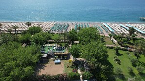 Vue aérienne d’un complexe hôtelier en bord de mer, avec des rangées de parasols et de chaises longues, un espace piscine et une végétation luxuriante.