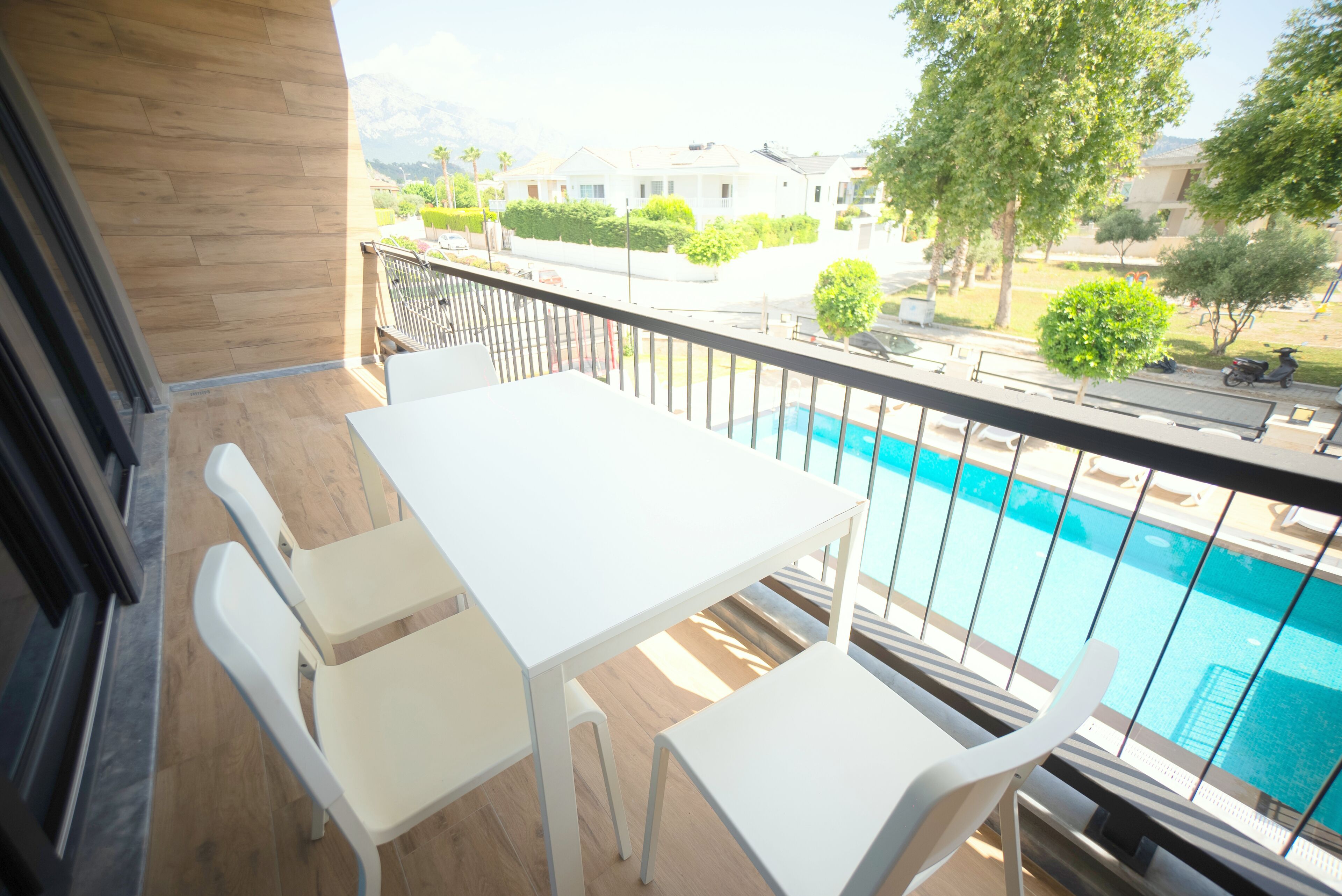 A balcony with a white table and chairs, overlooking a swimming pool and a residential area.