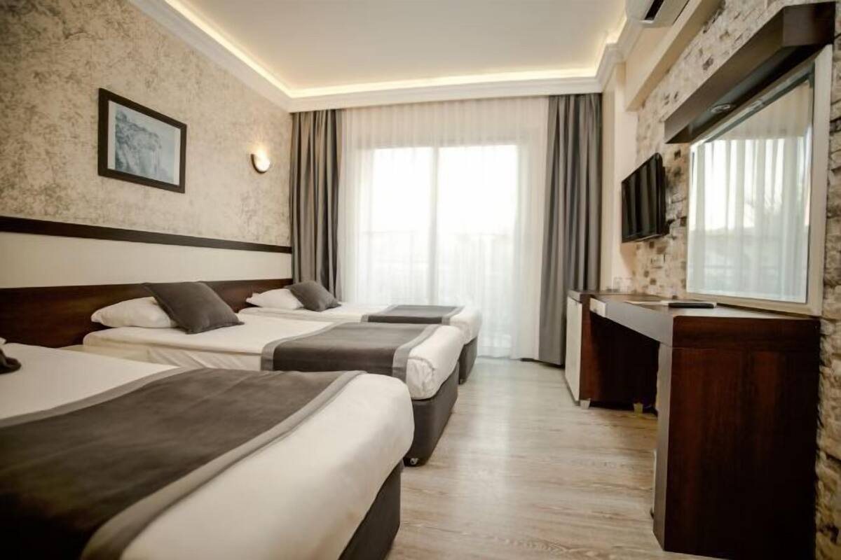 Une chambre d’hôtel avec trois lits, une télévision, un bureau et une grande fenêtre avec des rideaux.