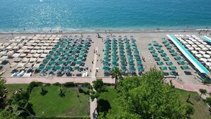 Une vue aérienne d’un complexe hôtelier en bord de mer, avec des rangées de parasols et de chaises longues, une vue dégagée sur l’océan et une pelouse bien entretenue.