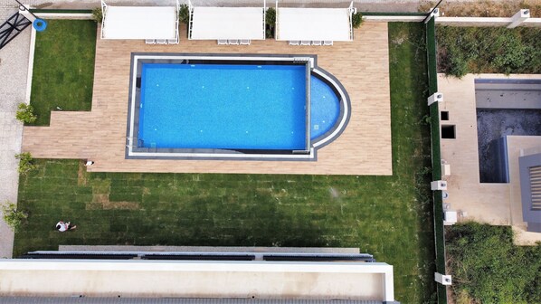 An aerial view of a swimming pool with a surrounding deck, a small garden area, and a building in the background.