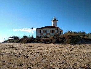 Un faro con fachada a cuadros y una torre alta, ubicado en una playa arenosa con vegetación escasa.