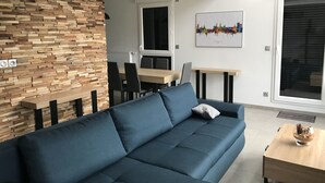 Ein modernes Wohnzimmer mit einem blauen Sofa, einer Wand mit Holzverkleidung und einem Essbereich mit Tisch und Stühlen.