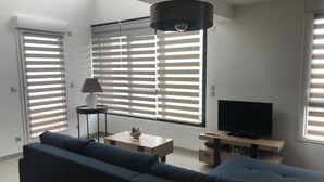 Ein modernes Wohnzimmer mit Sofa, Fernseher, Lampe und einem Fenster mit Jalousien.