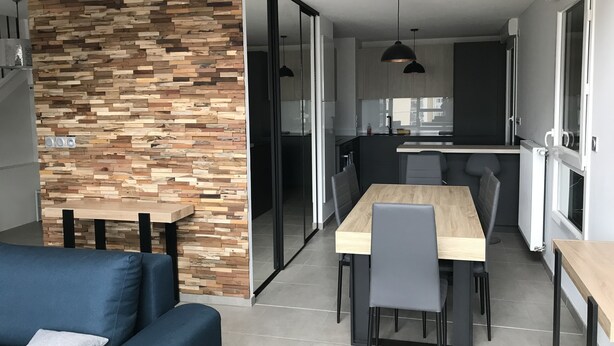 Eine moderne Küche mit einer integrierten Holzwand, einem Essbereich mit Holztisch und -stühlen sowie einem blauen Sofa mit Kissen.
