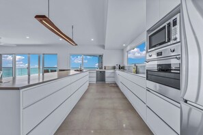 A modern kitchen with a central island, built-in appliances, and large windows offering a view of the sky. - The View (Airlie Beach)