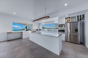 Une cuisine moderne dotée d’un grand îlot central, d’appareils électroménagers en acier inoxydable et offrant une vue sur la mer.