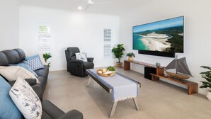 A modern living room with a sofa, armchair, coffee table, and a television displaying a scenic view. - The View (Airlie Beach)